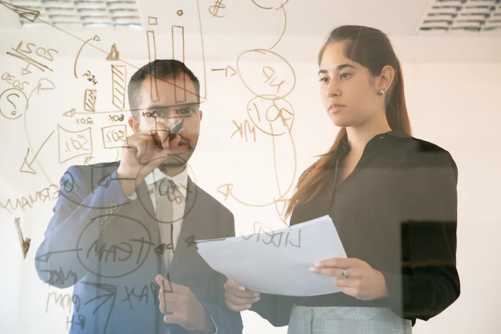 Two professionals, a man writing on a clear glass board covered with diagrams and a woman holding papers, engrossed in a discussion.