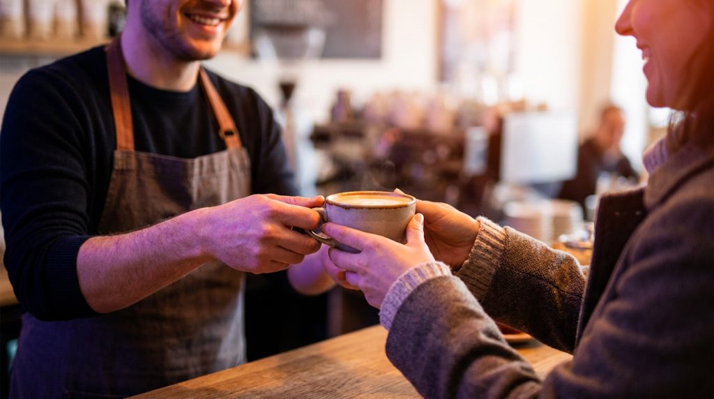 A barista handing a cup of coffee to a smiling customer across a counter.